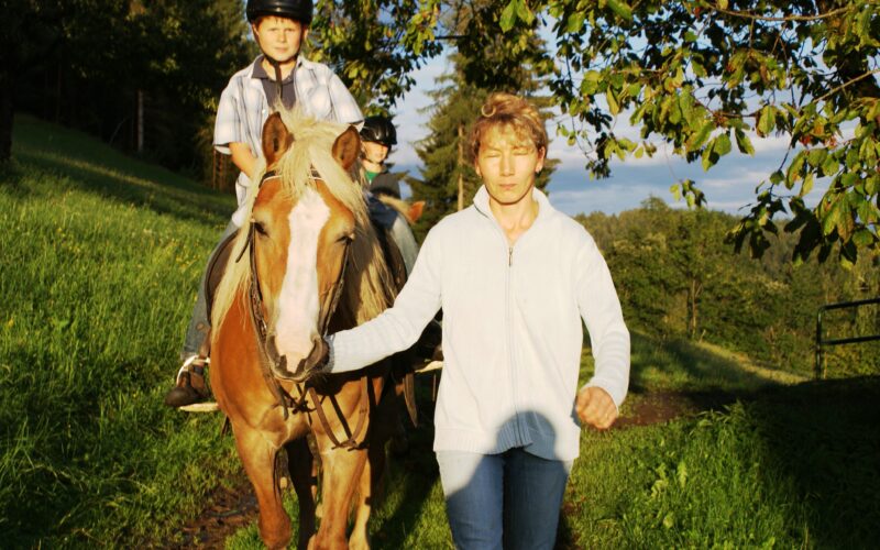 Eine Person führt ein hellbraunes Pferd mit blonder Mähne, während ein Kind mit einem Helm auf dem Rücken des Pferdes auf einer sonnenbeschienenen Wiese reitet.