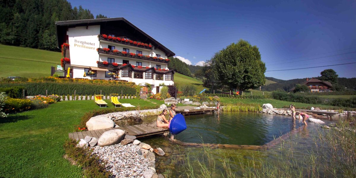 Ein Hotel mit blumengeschmückten Balkonen steht an einem natürlichen Schwimmteich, in dem sich die Menschen entspannen; grüne Wiesen und Hügel umgeben das Gebiet unter einem klaren blauen Himmel.
