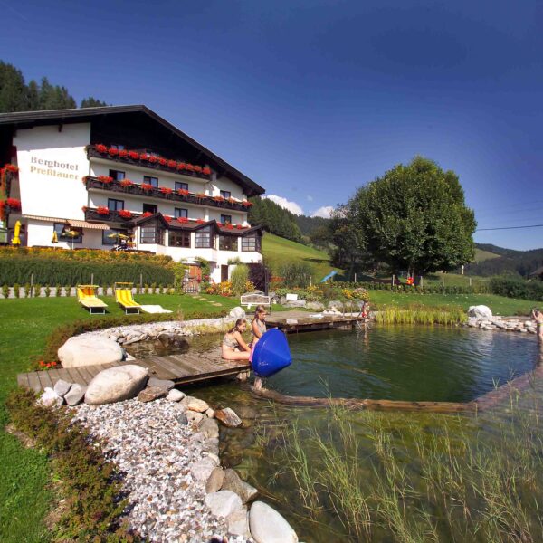 Ein Hotel mit blumengeschmückten Balkonen steht an einem natürlichen Schwimmteich, in dem sich die Menschen entspannen; grüne Wiesen und Hügel umgeben das Gebiet unter einem klaren blauen Himmel.