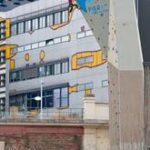 Eine Person klettert an einer künstlichen Kletterwand im Freien, mit einem modernen Gebäude mit bunten geometrischen Mustern im Hintergrund.