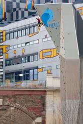 Eine Person klettert an einer künstlichen Kletterwand im Freien, mit einem modernen Gebäude mit bunten geometrischen Mustern im Hintergrund.