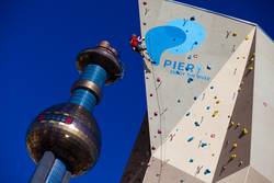 Eine Person klettert an einer künstlichen Kletterwand im Freien neben einem hohen, blau-silbernen Aussichtsturm vor einem klaren blauen Himmel.