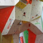 Eine Person klettert an einer großen Indoor-Kletterwand mit bunten Abschnitten und Griffen, umgeben von Bannern und natürlichem Licht aus Fenstern.