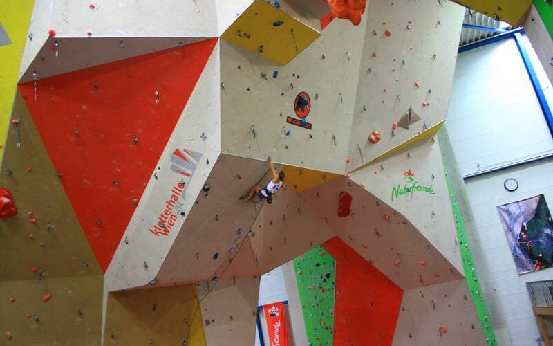 Eine Person klettert an einer großen Indoor-Kletterwand mit bunten Abschnitten und Griffen, umgeben von Bannern und natürlichem Licht aus Fenstern.
