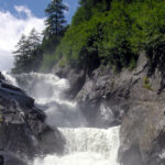 Ein mächtiger Wasserfall fließt über dunkle Felsklippen, umgeben von dichten grünen Bäumen und Vegetation unter einem teilweise bewölkten Himmel.