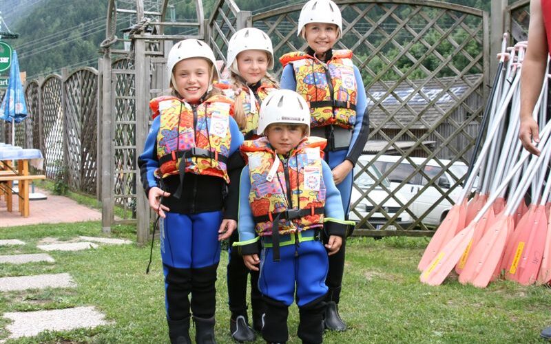Vier Kinder, die Neoprenanzüge, bunte Schwimmwesten und Helme tragen, stehen im Freien zusammen, während rechts die Paddel zu sehen sind.