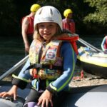 Ein junges Mädchen, das einen Helm und eine Schwimmweste trägt, sitzt auf einem aufblasbaren Floß an einem Fluss, mit anderen Menschen und Flößen im Hintergrund.