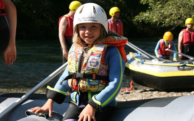 Ein junges Mädchen, das einen Helm und eine Schwimmweste trägt, sitzt auf einem aufblasbaren Floß an einem Fluss, mit anderen Menschen und Flößen im Hintergrund.