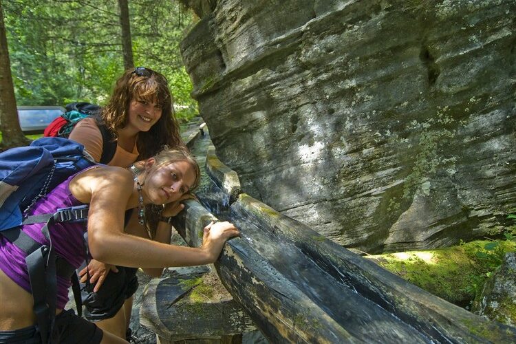 Zwei Frauen mit Rucksäcken lehnen sich über ein Holzgeländer neben einer Felsformation in einem bewaldeten Gebiet und scheinen aus einem hölzernen Wassertrog zu trinken.