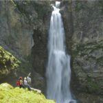 Vier Personen stehen auf einem grasbewachsenen Felsvorsprung in der Nähe des Fußes eines hohen, schmalen Wasserfalls, der eine felsige Klippe hinunterfließt.