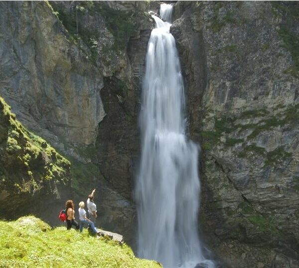 Vier Personen stehen auf einem grasbewachsenen Felsvorsprung in der Nähe des Fußes eines hohen, schmalen Wasserfalls, der eine felsige Klippe hinunterfließt.