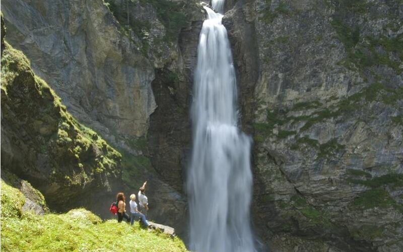 Vier Personen stehen auf einem grasbewachsenen Felsvorsprung in der Nähe des Fußes eines hohen, schmalen Wasserfalls, der eine felsige Klippe hinunterfließt.