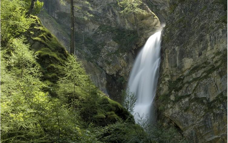 In einem Waldgebiet fließt ein Wasserfall eine felsige Klippe hinunter, umgeben von grünen Bäumen und moosbewachsenen Felsen.