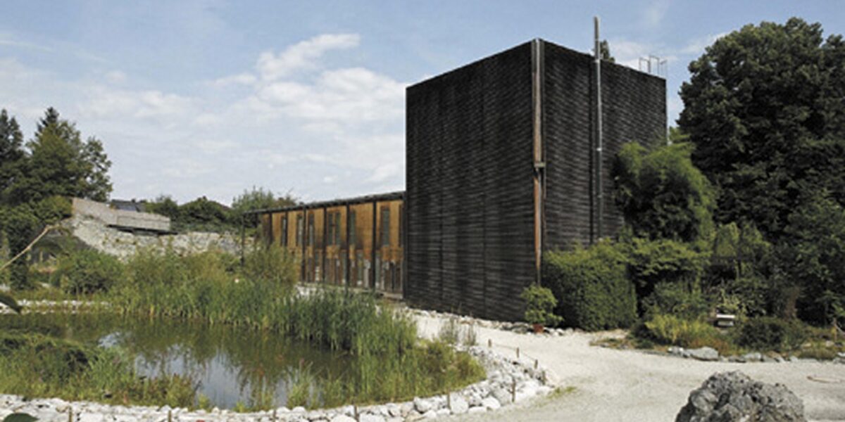 Ein modernes Gebäude mit dunkler Holzverkleidung steht neben einem Teich, umgeben von Kieswegen, Grünflächen und Bäumen unter einem teilweise bewölkten Himmel.