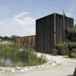 Ein modernes Gebäude mit dunkler Holzverkleidung steht neben einem Teich, umgeben von Kieswegen, Grünflächen und Bäumen unter einem teilweise bewölkten Himmel.