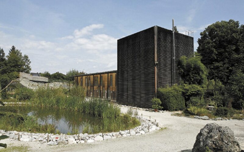 Ein modernes Gebäude mit dunkler Holzverkleidung steht neben einem Teich, umgeben von Kieswegen, Grünflächen und Bäumen unter einem teilweise bewölkten Himmel.