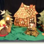 Drei Puppen und zwei Bäume sind vor einer Lebkuchenhaus-Kulisse auf einem grünen Tuch angeordnet, die eine Szene aus Hänsel und Gretel" darstellt.
