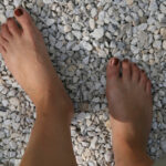 Zwei nackte Füße mit lackierten Zehennägeln stehen auf kleinen weißen Kieselsteinen im Freien, an deren Rändern etwas grünes Gras zu sehen ist.