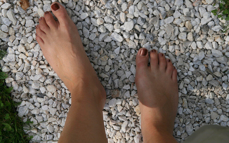 Zwei nackte Füße mit lackierten Zehennägeln stehen auf kleinen weißen Kieselsteinen im Freien, an deren Rändern etwas grünes Gras zu sehen ist.