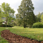 Ein grasbewachsener Park mit einem gewundenen Weg aus Holzspänen, umgeben von Bäumen und entfernten Häusern, mit Hügeln im Hintergrund unter einem bewölkten Himmel.