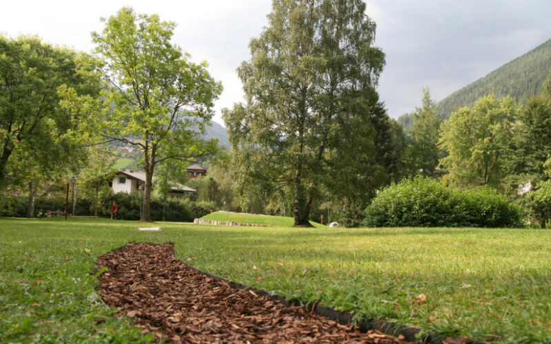 Ein grasbewachsener Park mit einem gewundenen Weg aus Holzspänen, umgeben von Bäumen und entfernten Häusern, mit Hügeln im Hintergrund unter einem bewölkten Himmel.