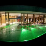 Ein modernes Gebäude mit großen Glasfenstern blickt auf einen nachts beleuchteten grünen Swimmingpool; im hell erleuchteten Innenraum sind Menschen zu sehen.