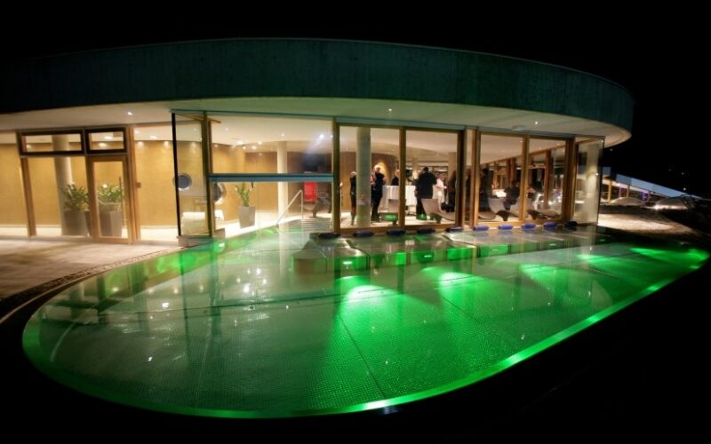 Ein modernes Gebäude mit großen Glasfenstern blickt auf einen nachts beleuchteten grünen Swimmingpool; im hell erleuchteten Innenraum sind Menschen zu sehen.