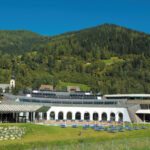 Ein moderner Ferienkomplex mit gewölbten Fenstern und Liegestühlen befindet sich vor einer Kulisse aus grünen Hügeln und bewaldeten Bergen unter einem klaren blauen Himmel.