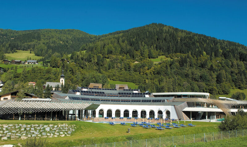 Ein moderner Ferienkomplex mit gewölbten Fenstern und Liegestühlen befindet sich vor einer Kulisse aus grünen Hügeln und bewaldeten Bergen unter einem klaren blauen Himmel.