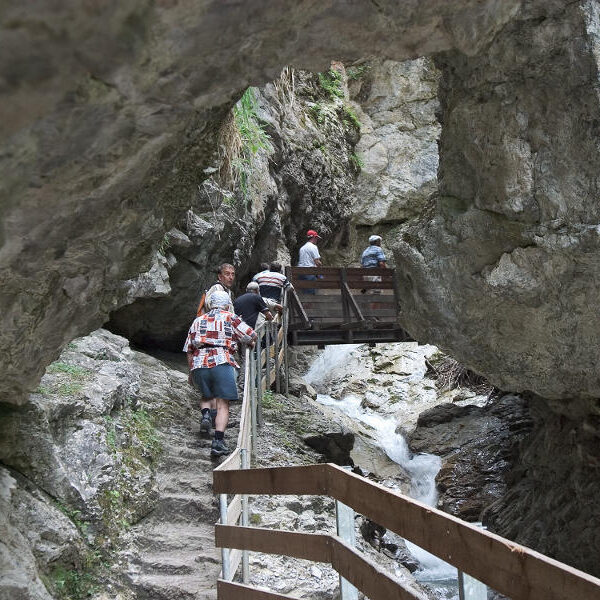 Die Menschen wandern über Steinstufen und eine schmale Holzbrücke in einer felsigen Schlucht mit einem kleinen Bach darunter.