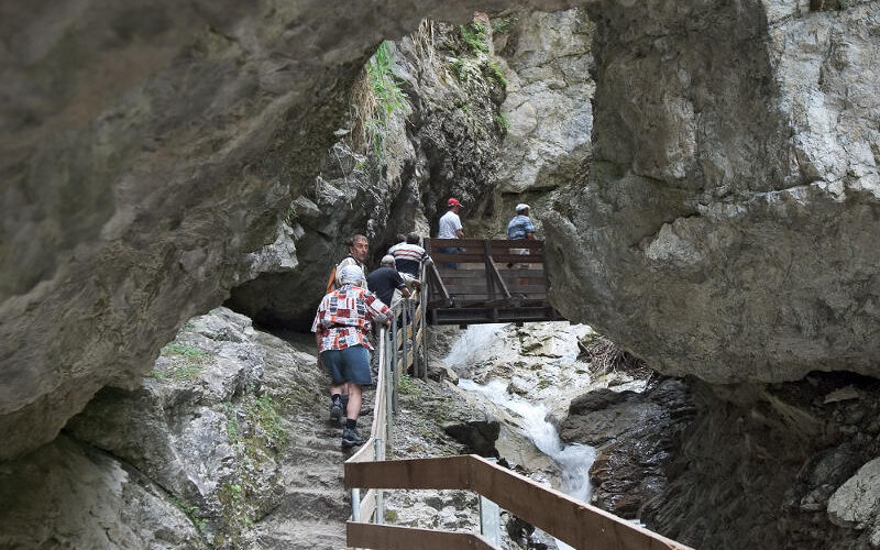 Die Menschen wandern über Steinstufen und eine schmale Holzbrücke in einer felsigen Schlucht mit einem kleinen Bach darunter.