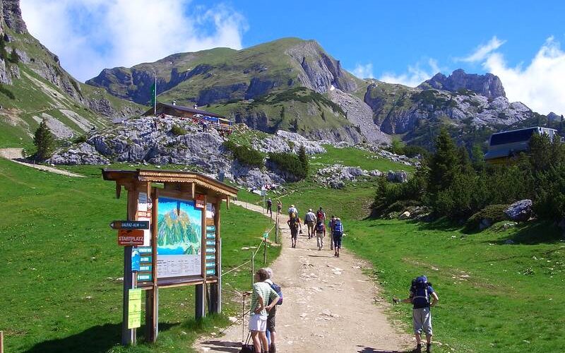 Wanderer gehen einen breiten unbefestigten Weg in einer bergigen Landschaft hinauf, mit einem großen Schild im Vordergrund und grünen Hügeln und felsigen Gipfeln im Hintergrund.