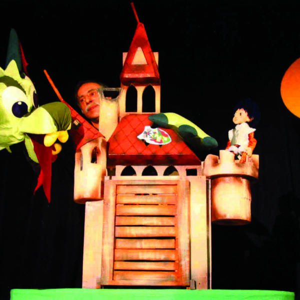 Ein Puppenspieler bedient eine grüne Drachenpuppe und ein Schlossrequisit auf der Bühne, während im Hintergrund eine kleine Puppenfigur und ein orangefarbener Kreis, der der Sonne ähnelt, zu sehen sind.