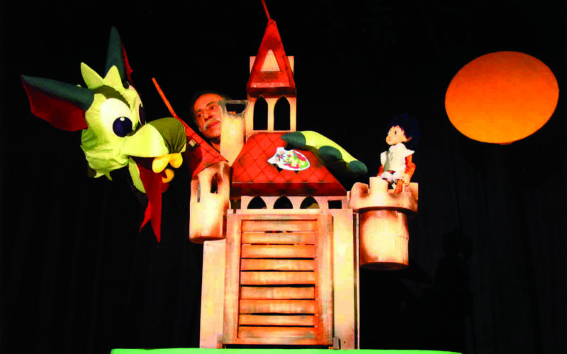 Ein Puppenspieler bedient eine grüne Drachenpuppe und ein Schlossrequisit auf der Bühne, während im Hintergrund eine kleine Puppenfigur und ein orangefarbener Kreis, der der Sonne ähnelt, zu sehen sind.