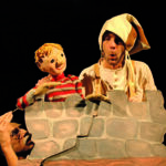 Ein Darsteller mit Narrenhut hält eine gestreifte Puppe hinter einer bemalten Steinwand, während eine andere Person von unten heraufschaut.