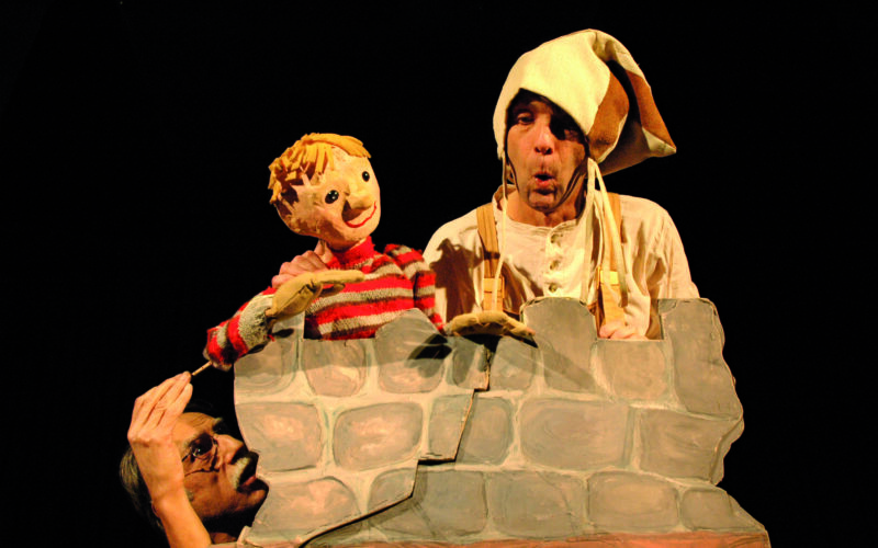 Ein Darsteller mit Narrenhut hält eine gestreifte Puppe hinter einer bemalten Steinwand, während eine andere Person von unten heraufschaut.