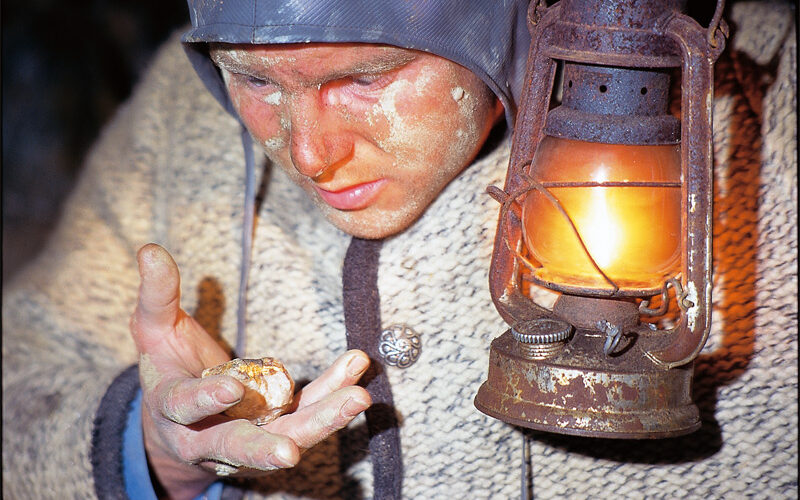 Eine Person mit Helm und warmer Kleidung untersucht im Licht einer Laterne einen Goldnugget, wobei Gesicht und Hände mit Staub bedeckt sind.