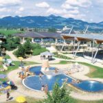 Außenpoolkomplex mit Menschen, die sich sonnen und schwimmen, umgeben von Sonnenschirmen, Gras und Gebäuden, im Hintergrund sind Berge zu sehen.