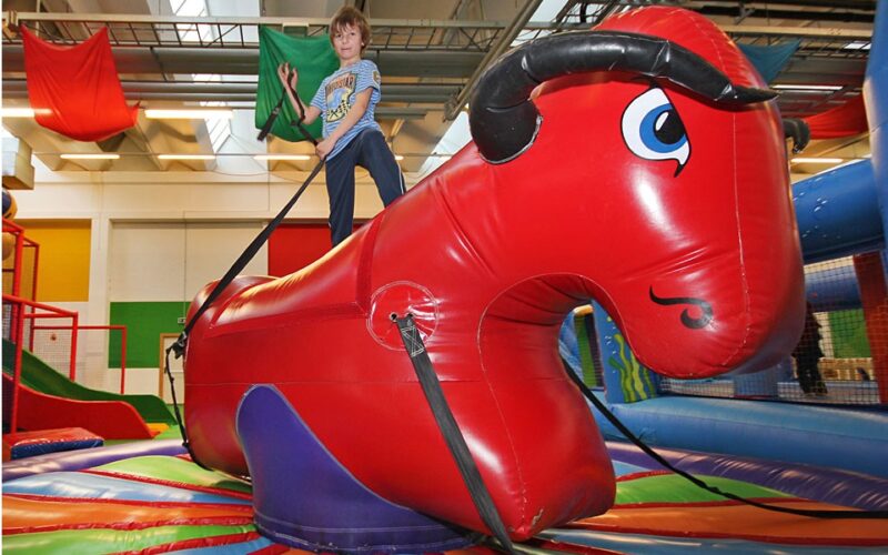 Ein Kind reitet auf einem roten, aufblasbaren mechanischen Stier in einem bunt geschmückten Innenspielbereich.