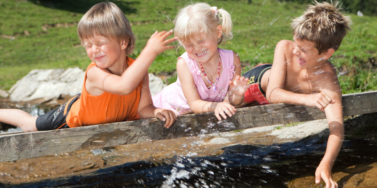 Drei kleine Kinder lehnen sich an einem sonnigen Tag mit grünem Gras im Hintergrund lachend und spritzend über einen Holzvorsprung an einem Bach.