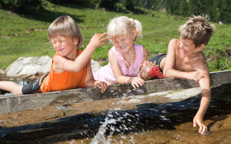 Drei kleine Kinder lehnen sich an einem sonnigen Tag mit grünem Gras im Hintergrund lachend und spritzend über einen Holzvorsprung an einem Bach.