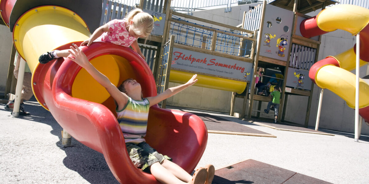 Zwei Kinder spielen auf einer rot-gelben Rutsche auf einem Spielplatz im Freien, wobei eines am unteren Ende der Rutsche sitzt und das andere sich darauf vorbereitet, hinunterzurutschen.