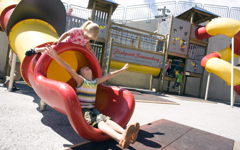 Zwei Kinder spielen auf einer rot-gelben Rutsche auf einem Spielplatz im Freien, wobei eines am unteren Ende der Rutsche sitzt und das andere sich darauf vorbereitet, hinunterzurutschen.