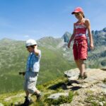 Zwei Kinder wandern auf einem felsigen Bergpfad mit grünen Hügeln und zerklüfteten Gipfeln im Hintergrund unter einem klaren blauen Himmel.