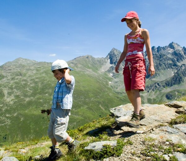 Zwei Kinder wandern auf einem felsigen Bergpfad mit grünen Hügeln und zerklüfteten Gipfeln im Hintergrund unter einem klaren blauen Himmel.