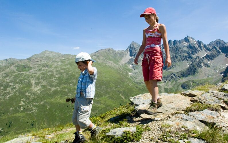 Zwei Kinder wandern auf einem felsigen Bergpfad mit grünen Hügeln und zerklüfteten Gipfeln im Hintergrund unter einem klaren blauen Himmel.
