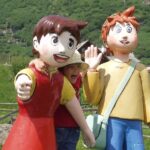 Ein Kind posiert mit zwei großen Zeichentrickfiguren im Freien, mit Grünflächen und Holzgebäuden im Hintergrund.