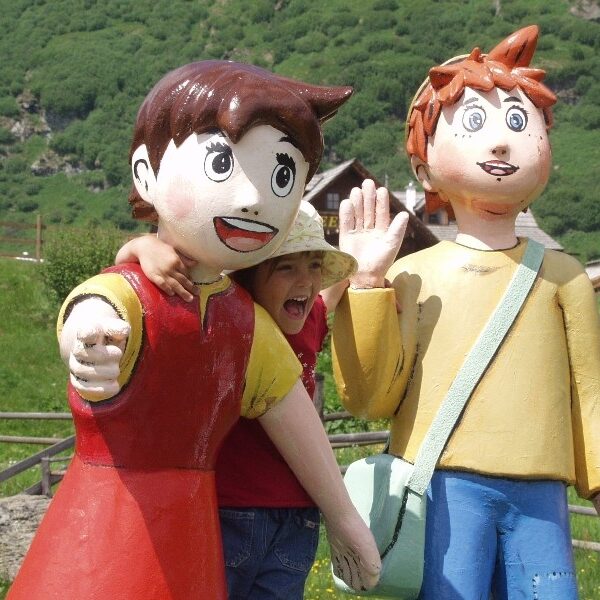 Ein Kind posiert mit zwei großen Zeichentrickfiguren im Freien, mit Grünflächen und Holzgebäuden im Hintergrund.