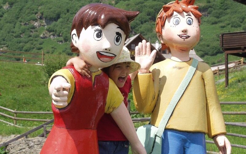 Ein Kind posiert mit zwei großen Zeichentrickfiguren im Freien, mit Grünflächen und Holzgebäuden im Hintergrund.