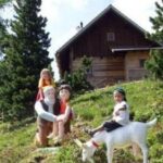 Drei Kinder posieren neben lebensgroßen Statuen von Figuren und einer Ziege auf einem grasbewachsenen Hügel mit einer Holzhütte und Bäumen im Hintergrund.